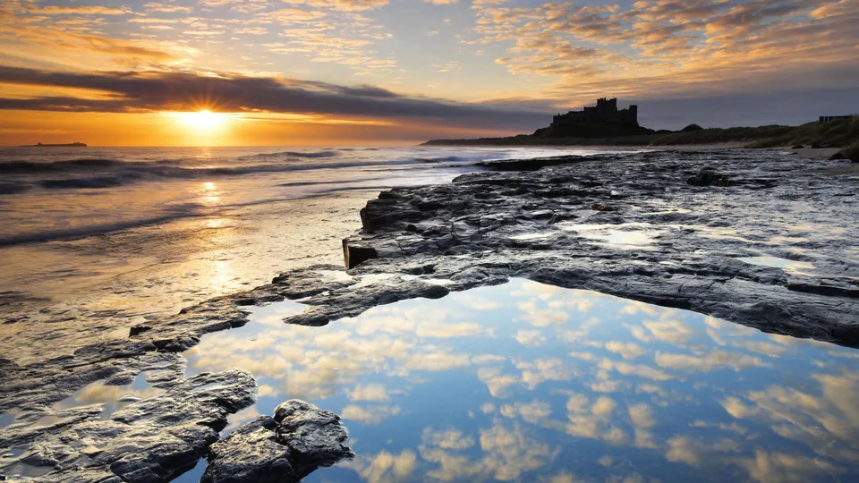 A dramatic sunset over a rocky coastline with a castle in the distance and a reflection in a tidal pool.