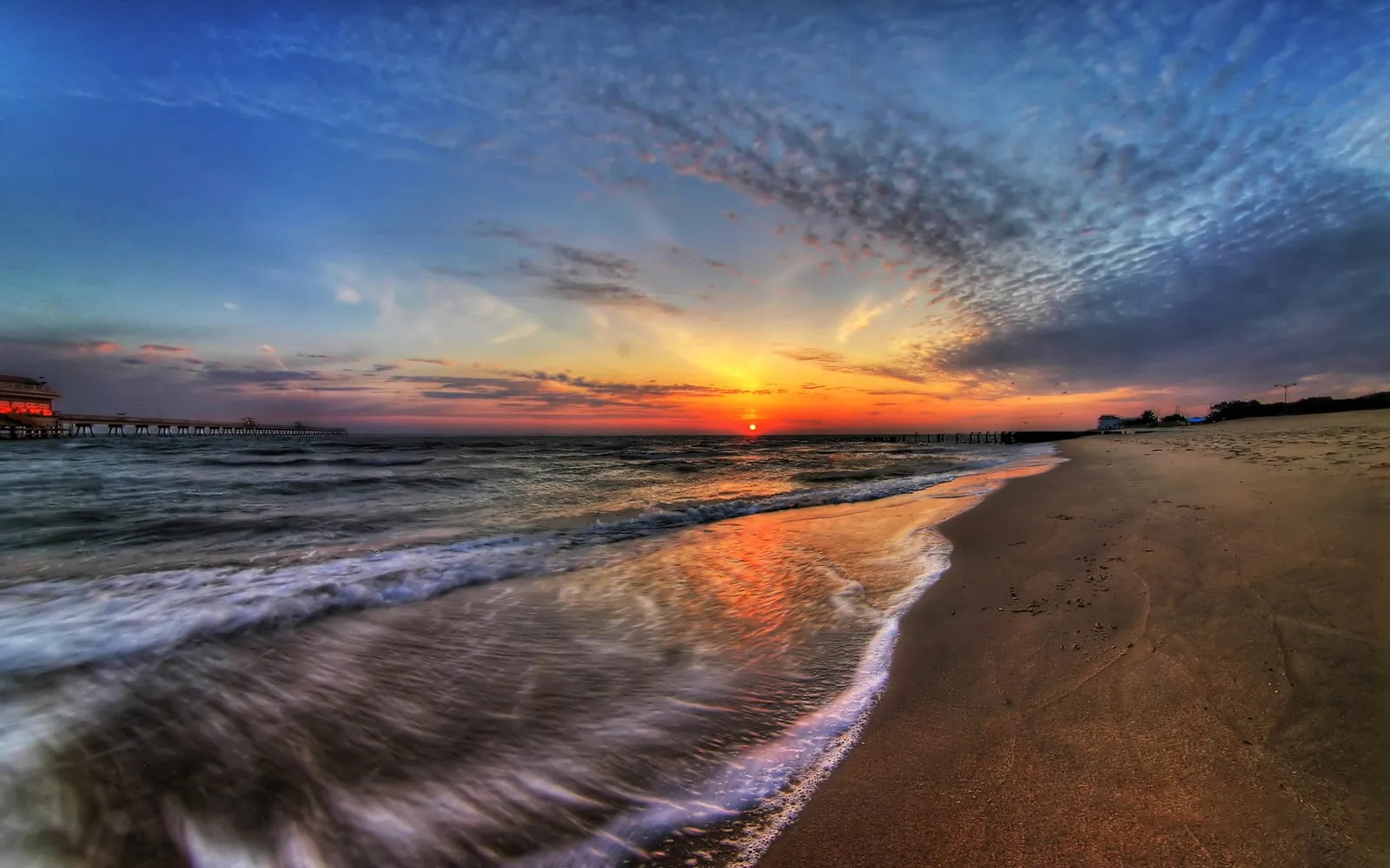 The sun sets over the ocean, reflecting on the wet sand and waves.