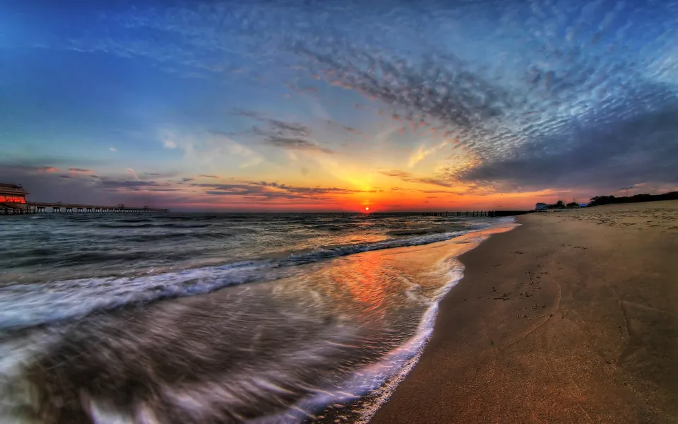 The sun sets over the ocean, reflecting on the wet sand and waves.
