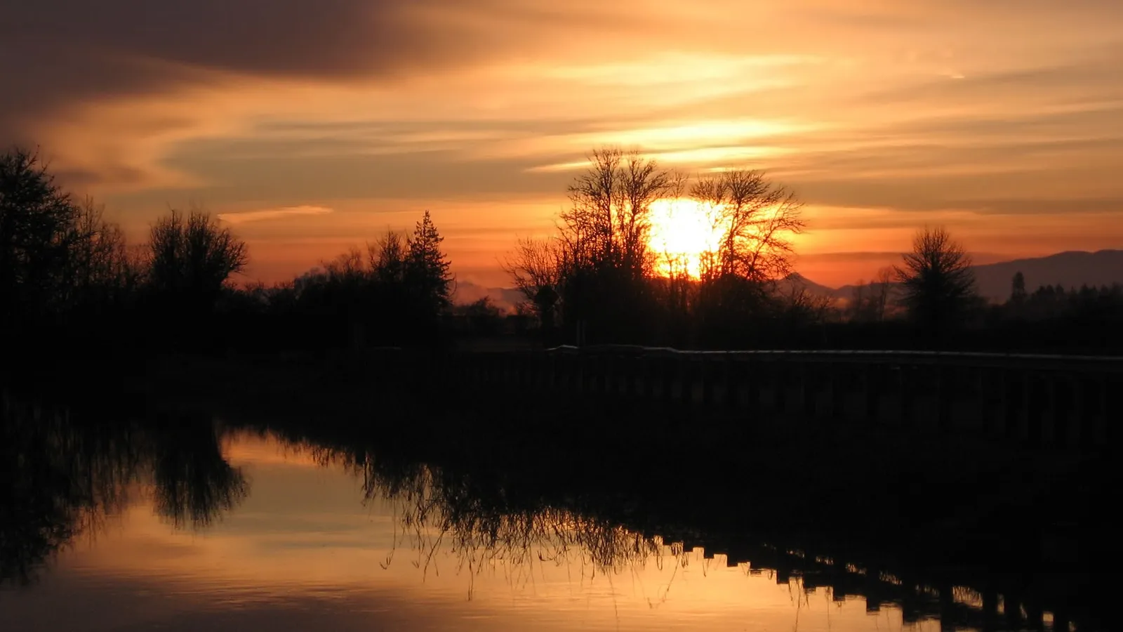 A vibrant sunset with orange and yellow hues reflects in the still water, with silhouetted trees and a distant mountain range.