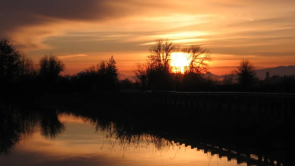 A vibrant sunset with orange and yellow hues reflects in the still water, with silhouetted trees and a distant mountain range.