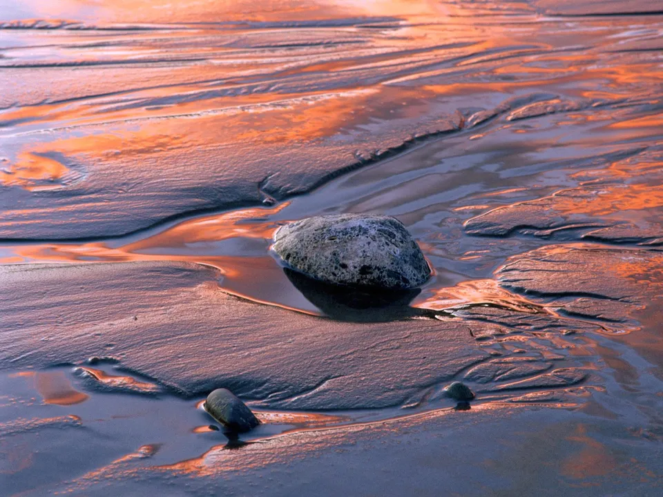 Close-up of wet sand with ripples, reflecting orange sunset light, with rocks in tidal pools.