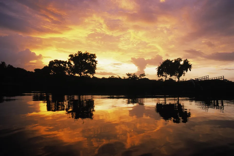 A vibrant sunset with orange and yellow clouds reflects in the calm waters of a river, silhouetting trees and a small structure.