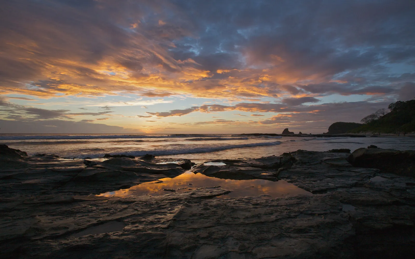 A dramatic sunset over a rocky coastline with waves crashing and colorful clouds reflected in tide pools.