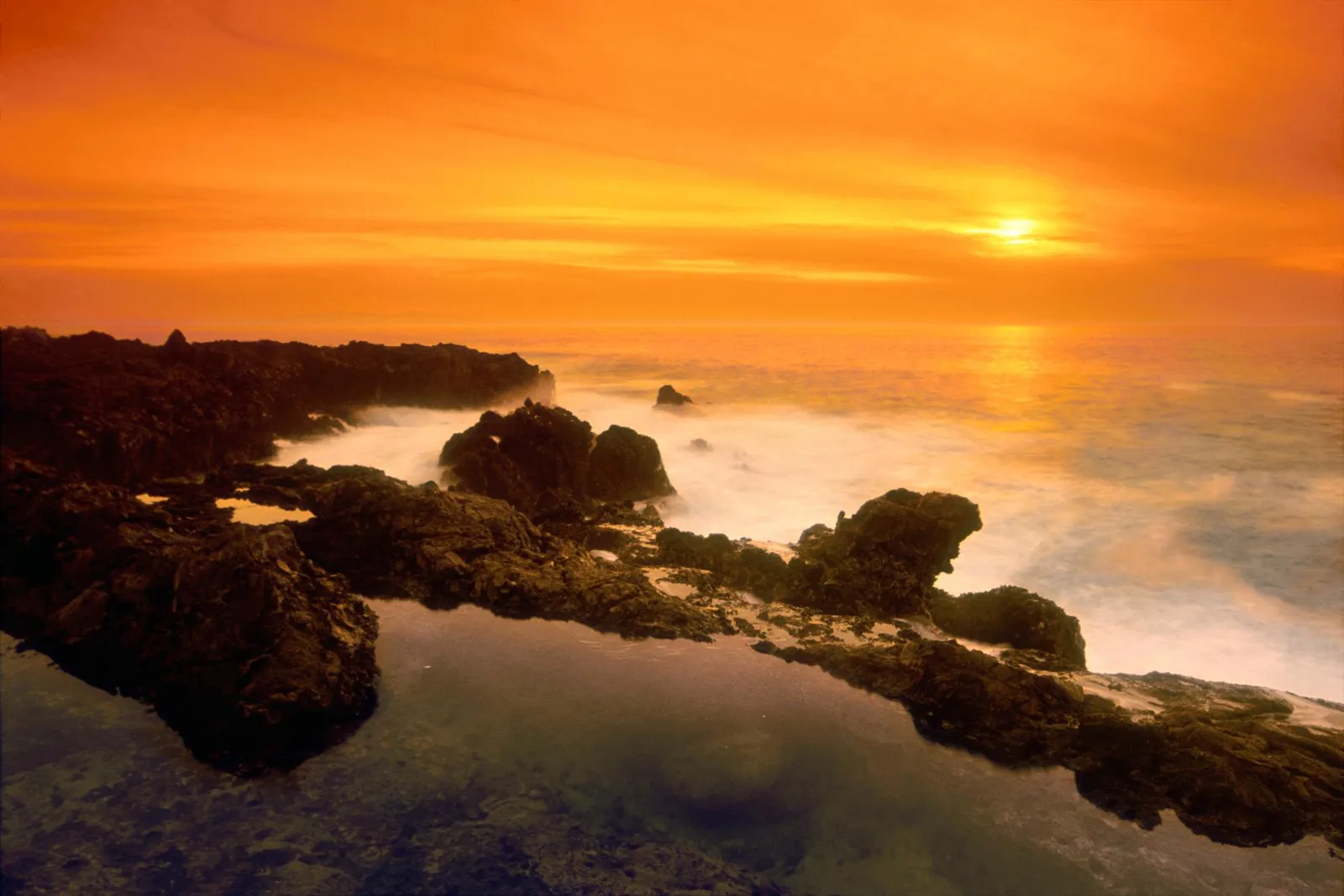 A dramatic sunset with vibrant orange and yellow hues over a rocky coastline and the ocean.