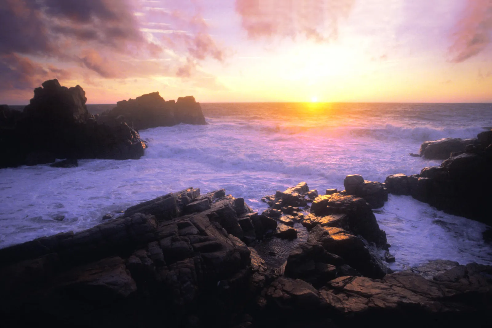 A vibrant sunset casts a warm glow over a rocky coastline with waves crashing against the shore.