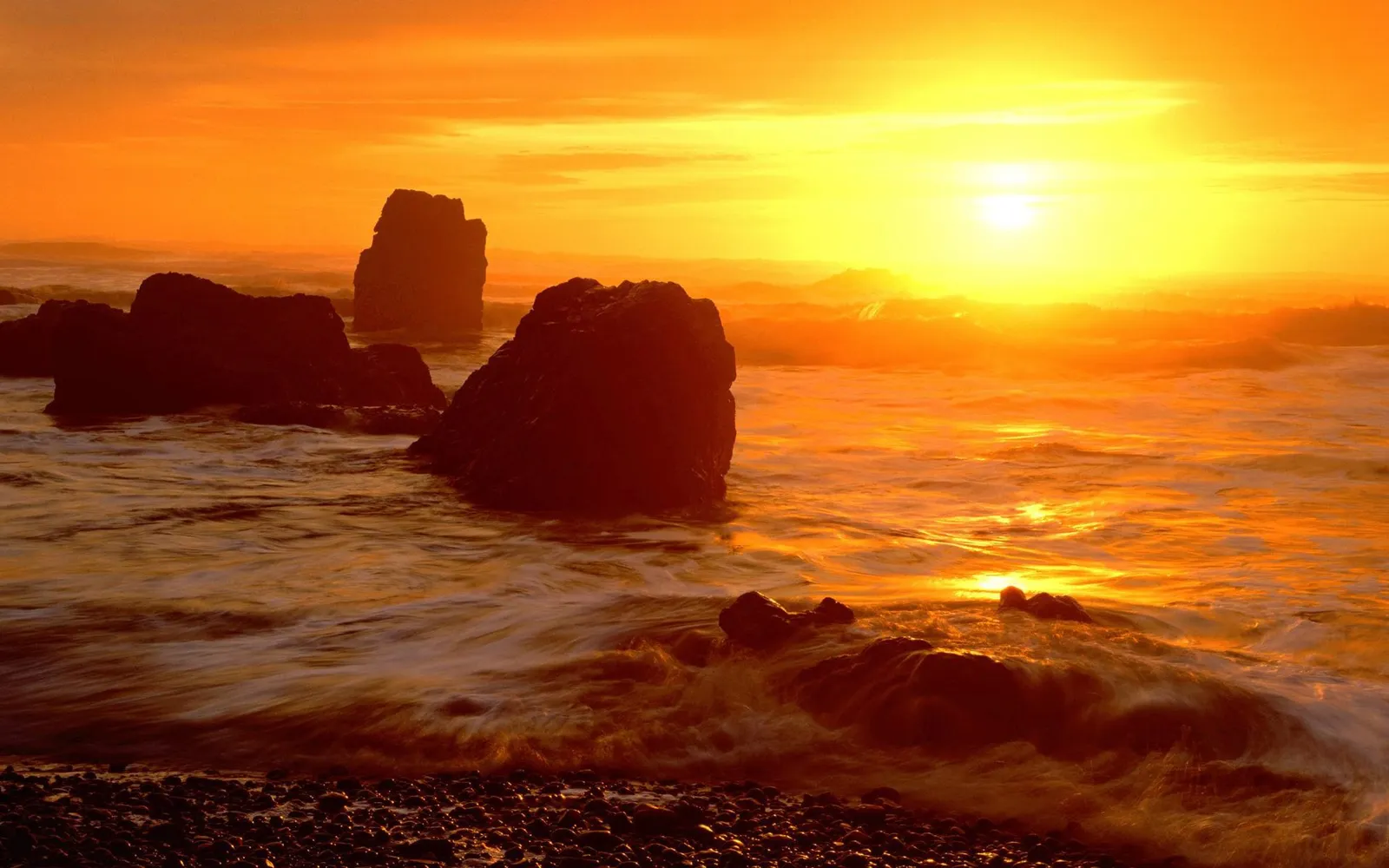 A vibrant orange sunset illuminates a rocky coastline with waves crashing against dark sea stacks.