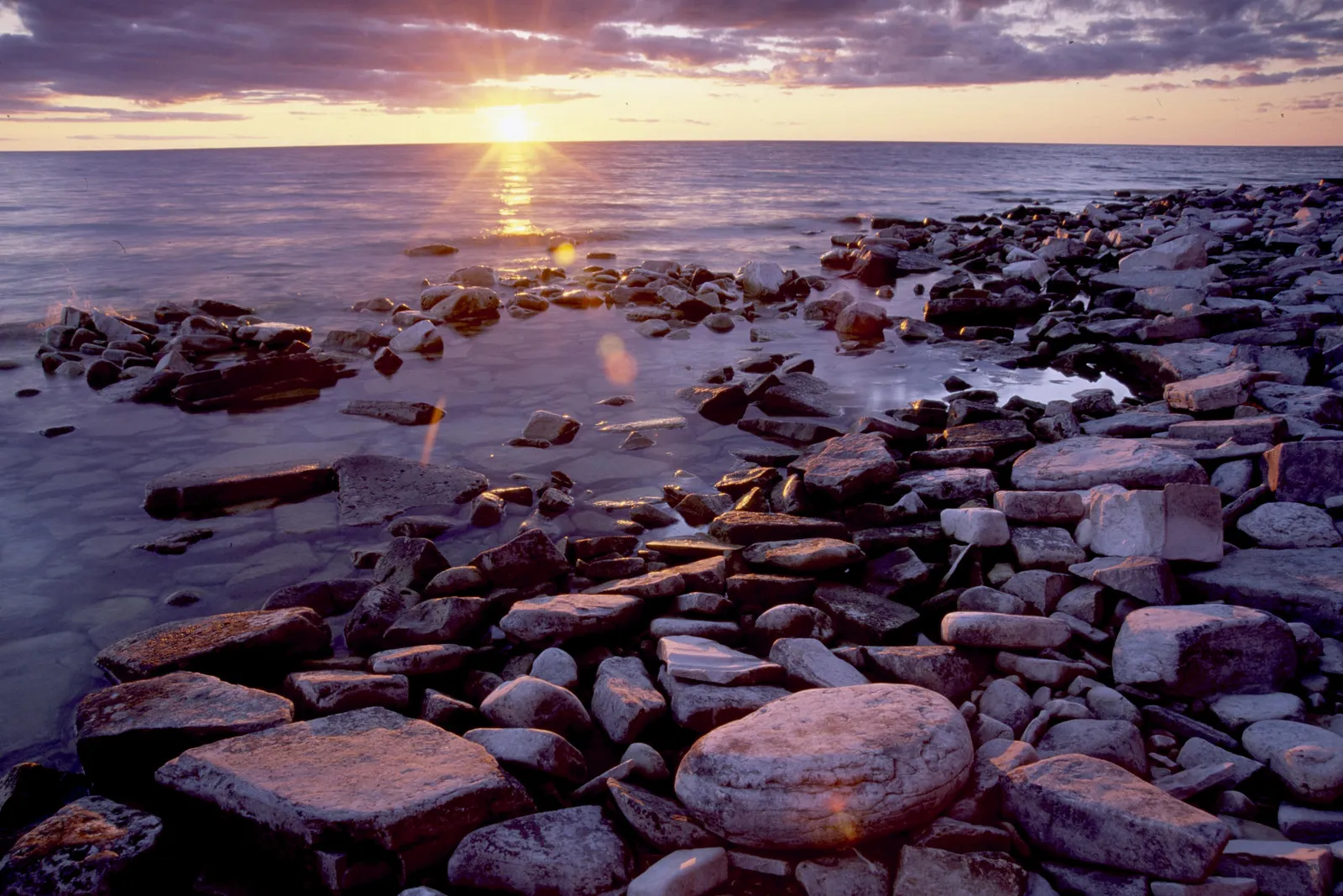 A vibrant sunset casts golden light over a rocky shoreline and calm water.
