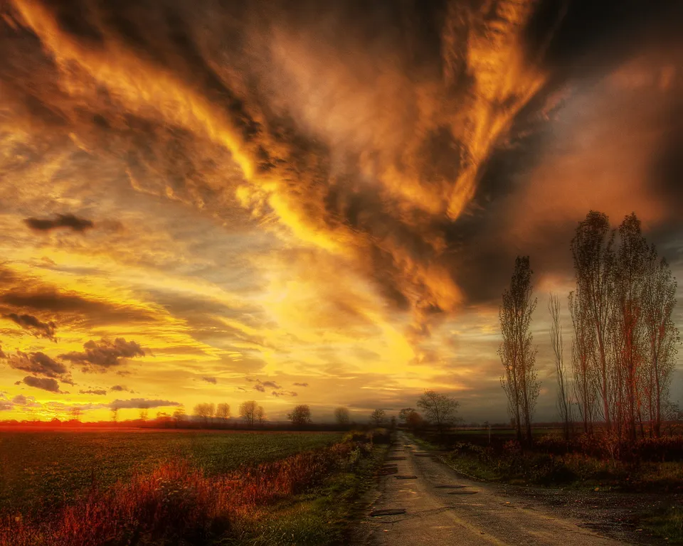A dramatic sunset with fiery orange and yellow clouds over a rural road and fields.