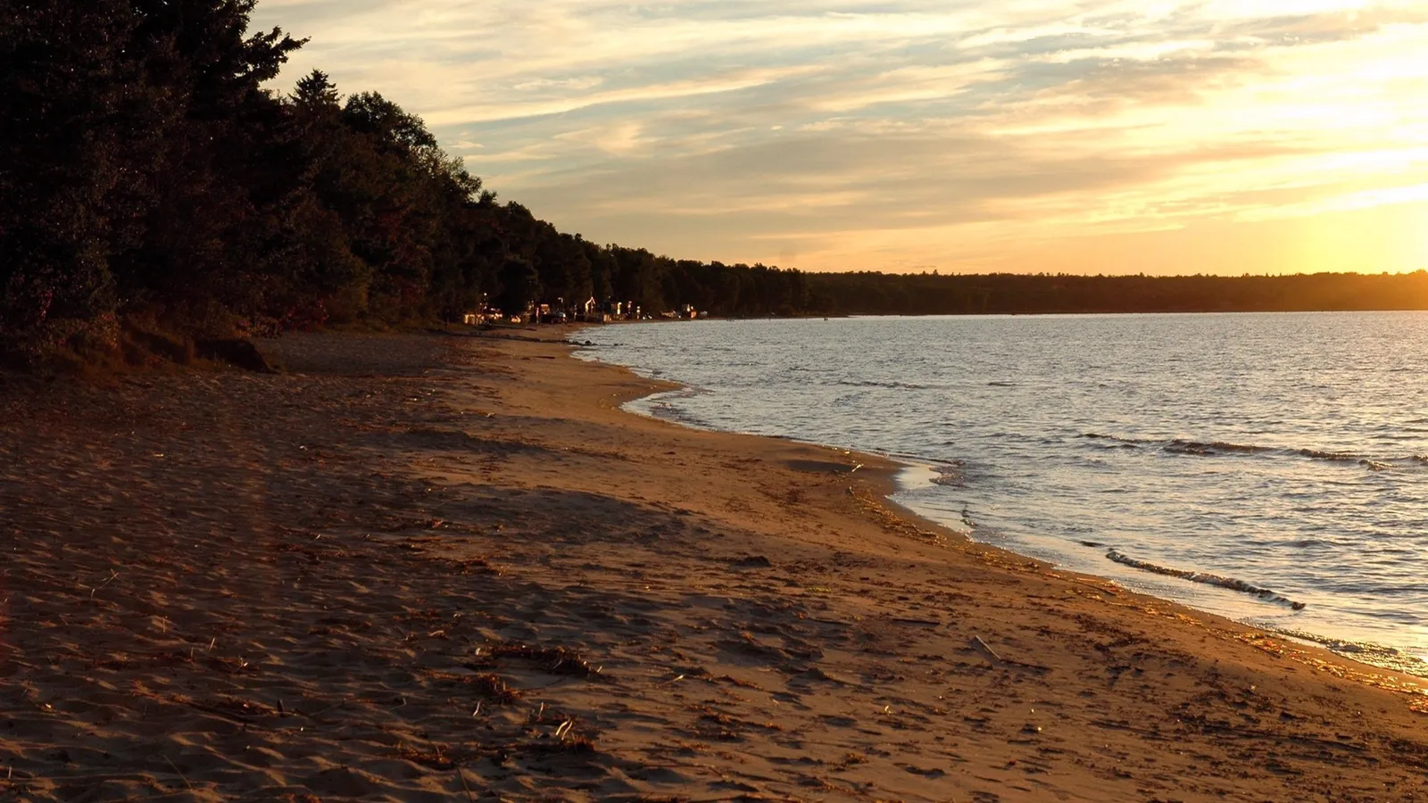 A sandy beach meets calm water under a golden sunset sky with wispy clouds.