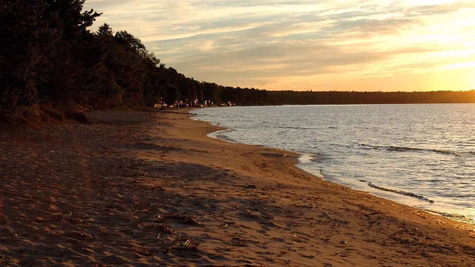 A sandy beach meets calm water under a golden sunset sky with wispy clouds.
