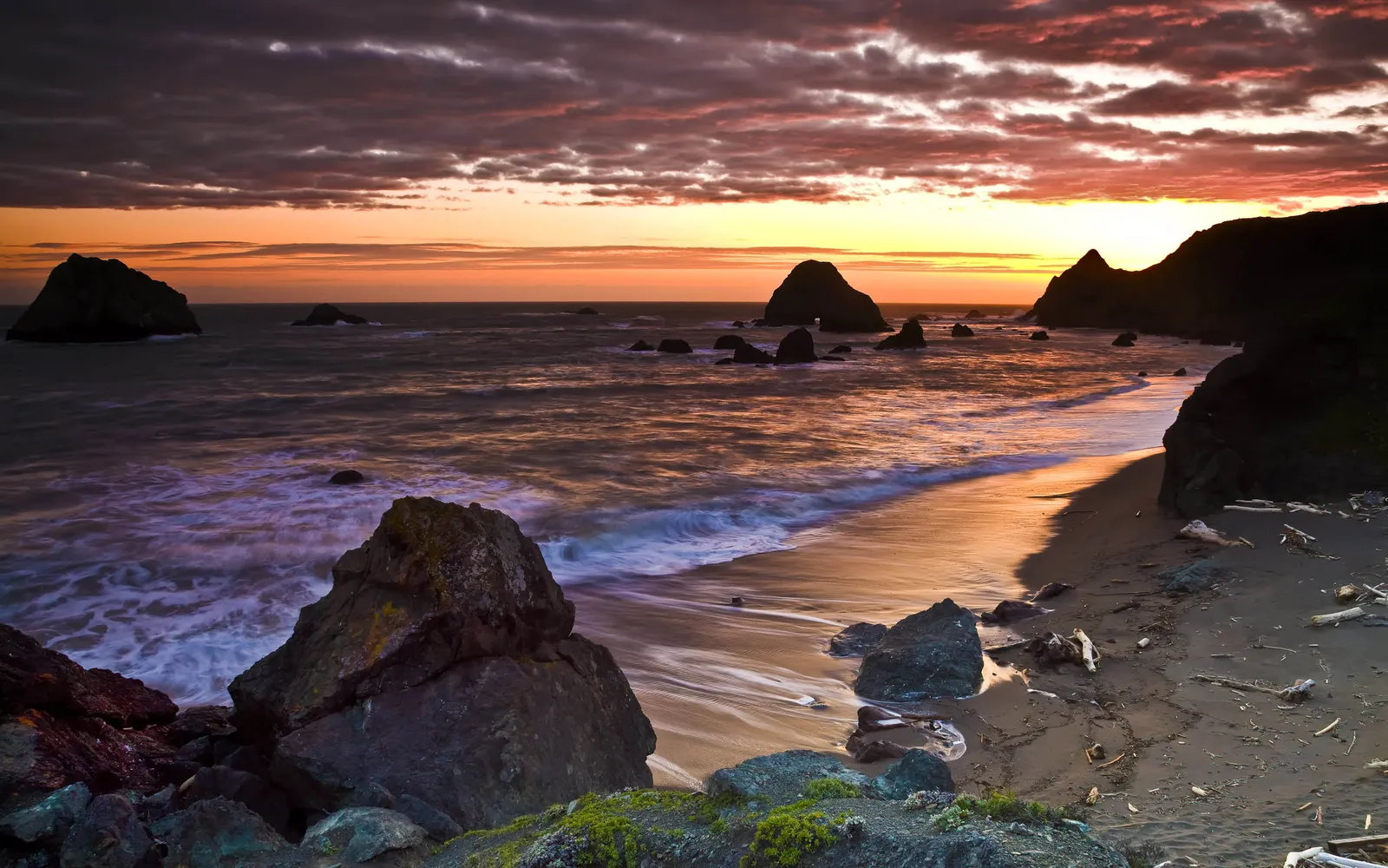 The sun sets over the ocean, casting a warm glow on a sandy beach with waves lapping against dark rocks.
