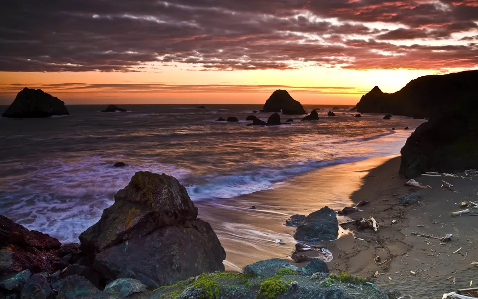 The sun sets over the ocean, casting a warm glow on a sandy beach with waves lapping against dark rocks.
