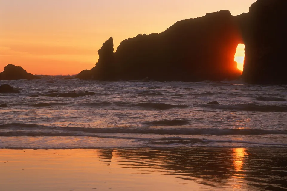 The sun sets through a natural archway in a rocky cliff face overlooking the ocean.
