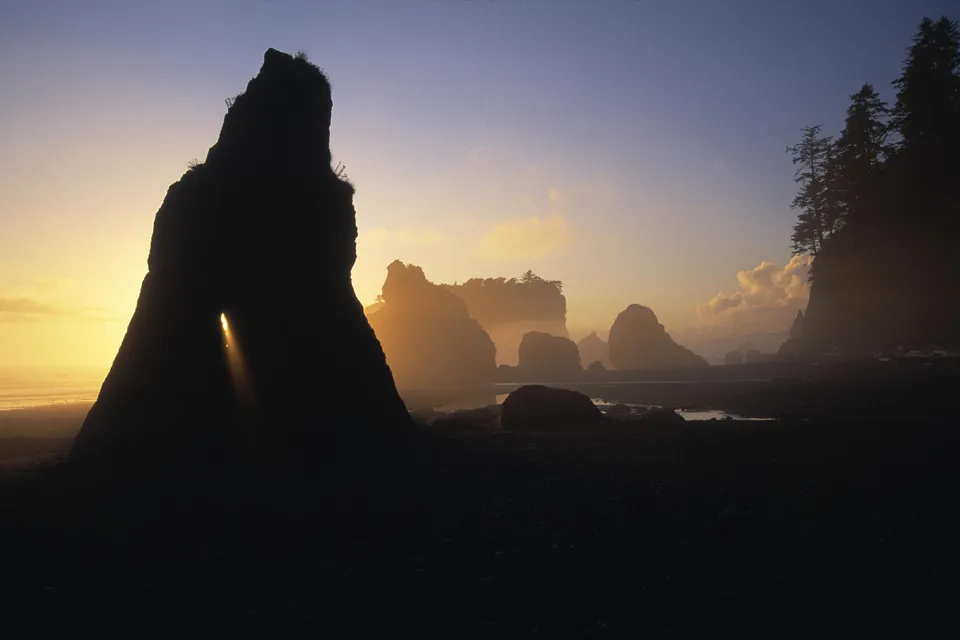 A dramatic sea stack with an arch is silhouetted against a golden sunset sky, with other sea stacks in the background.