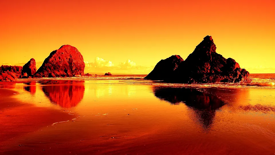 Silhouetted sea stacks stand against a vibrant orange and yellow sunset sky reflected on a wet sandy beach.