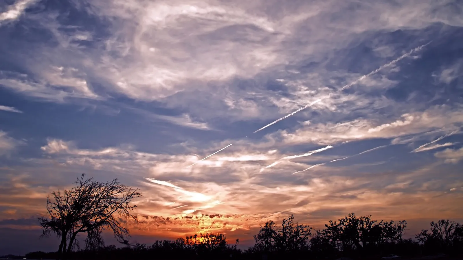 A vibrant sunset sky with orange and pink clouds, streaked with white contrails from airplanes.