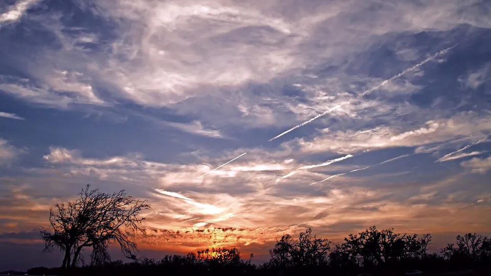 A vibrant sunset sky with orange and pink clouds, streaked with white contrails from airplanes.