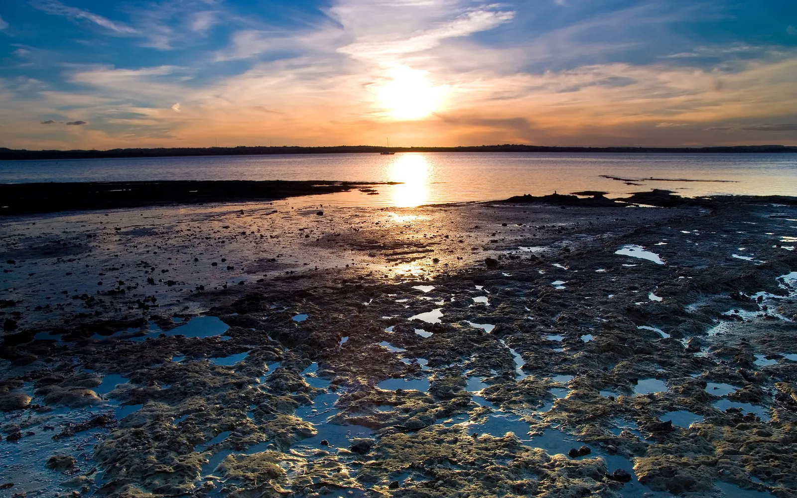 A vibrant sunset over a calm body of water, with a rocky, wet tidal flat in the foreground reflecting the sky.