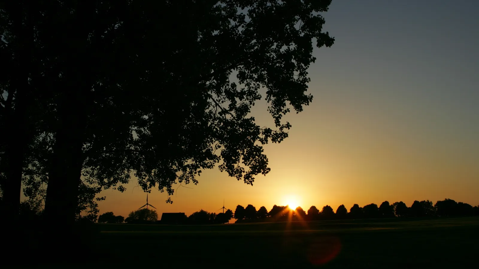 A silhouette of trees and a wind turbine against a bright orange and yellow sunset.