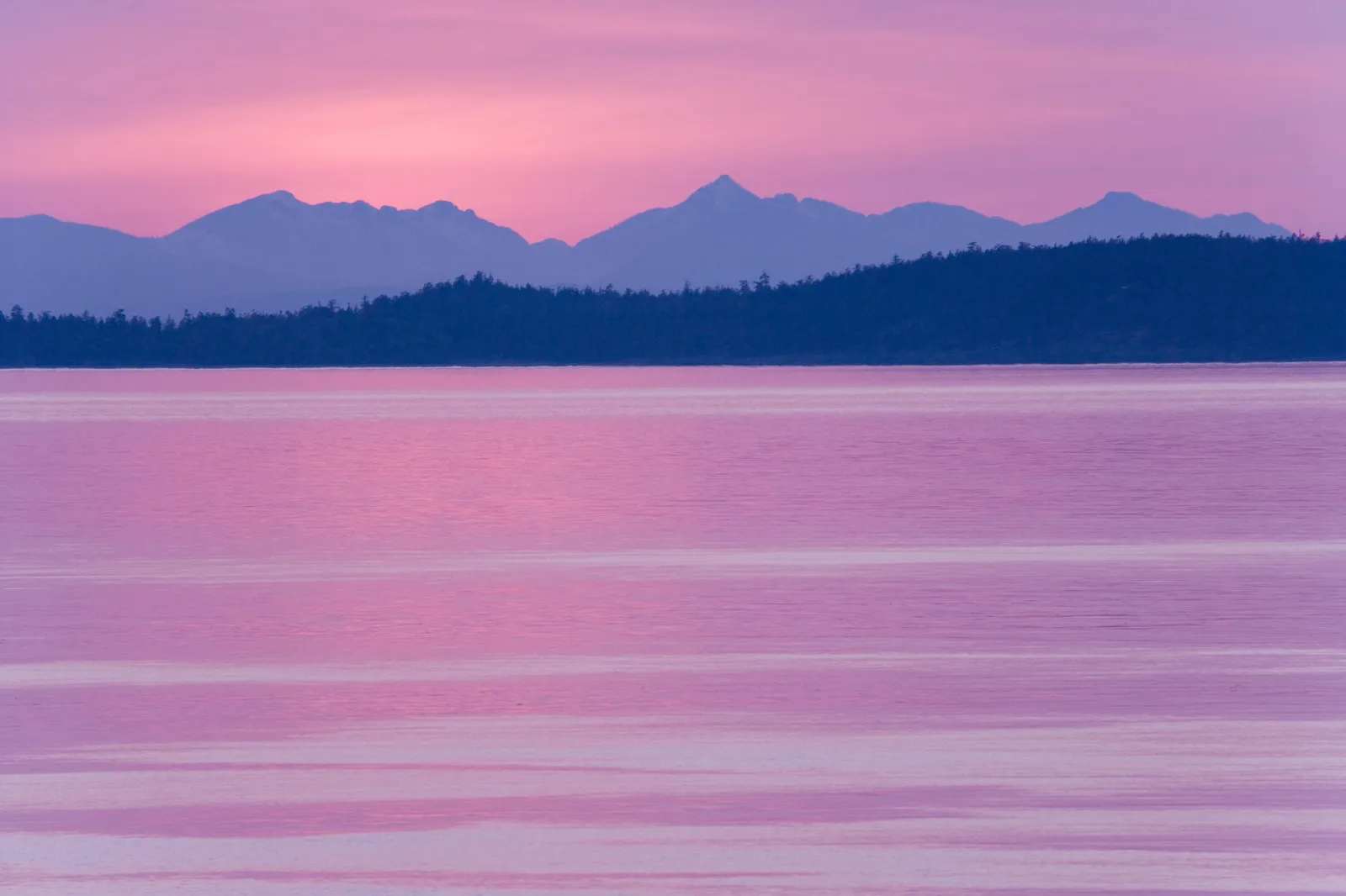 A tranquil body of water reflects the pink and purple hues of a sunset sky behind silhouetted mountains and trees.