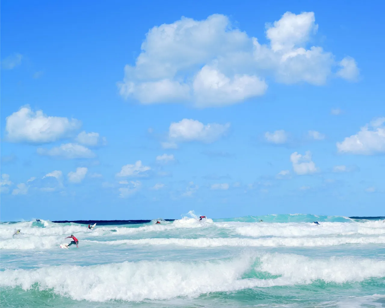 Several surfers are riding waves on a bright, sunny day with a blue sky and scattered clouds.