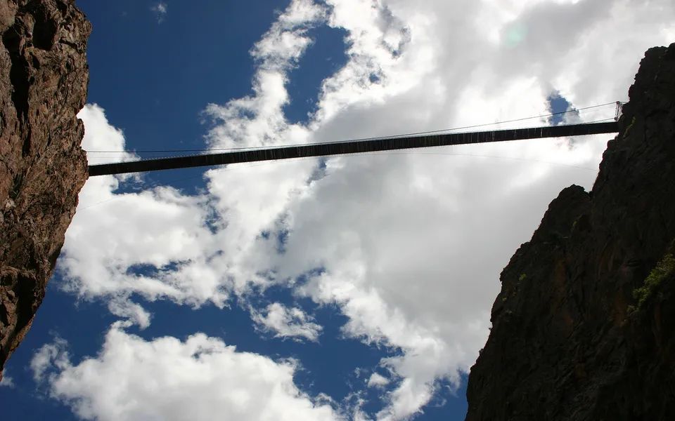 A long suspension bridge stretches across a deep canyon, with dramatic clouds in the blue sky above.