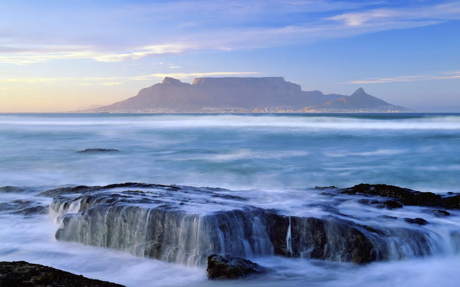 A long exposure shot of waves crashing on rocks with Table Mountain in the background.