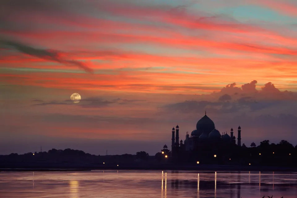 The Taj Mahal is silhouetted against a vibrant sunset sky with a full moon visible.