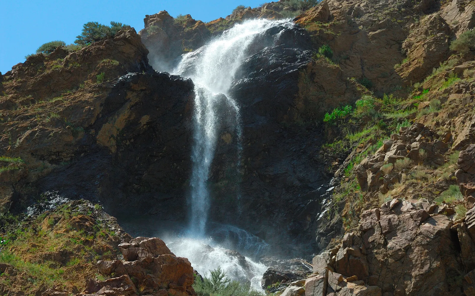 A tall waterfall cascades down a dark, rugged cliff face with sparse vegetation.