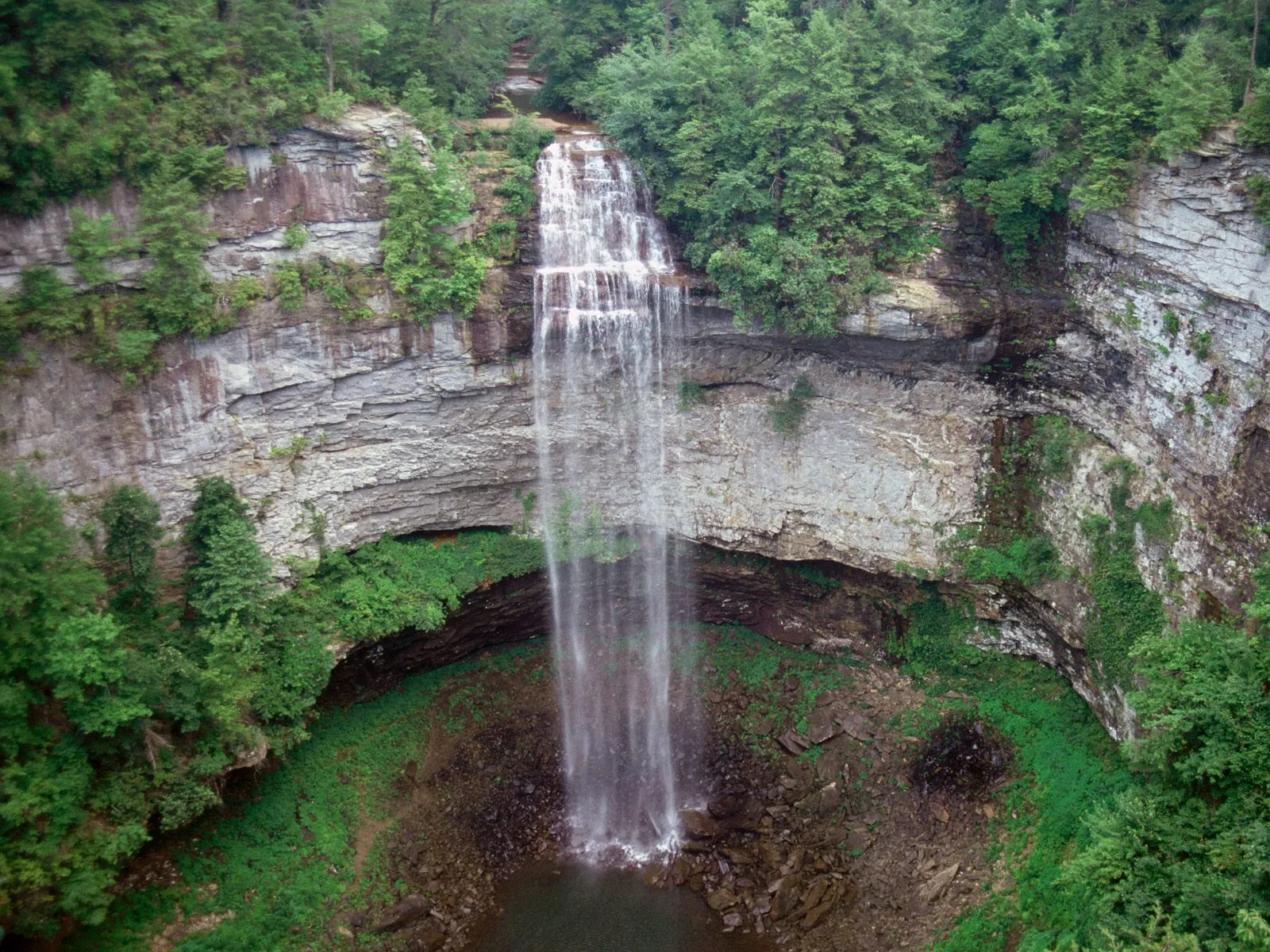 A tall, thin waterfall plunges from a high, layered rock cliff surrounded by lush green trees.