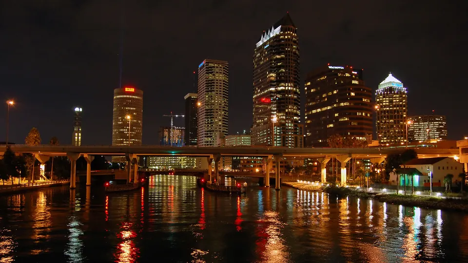 A nighttime cityscape of Tampa, Florida, with illuminated skyscrapers reflected in the dark water.
