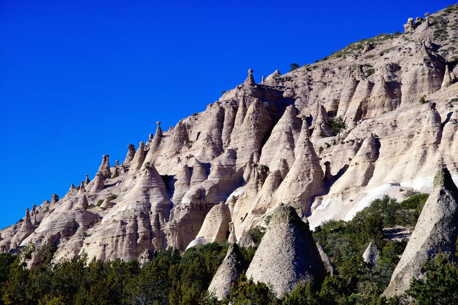 Cone-shaped rock formations called hoodoos rise up a hillside under a bright blue sky.