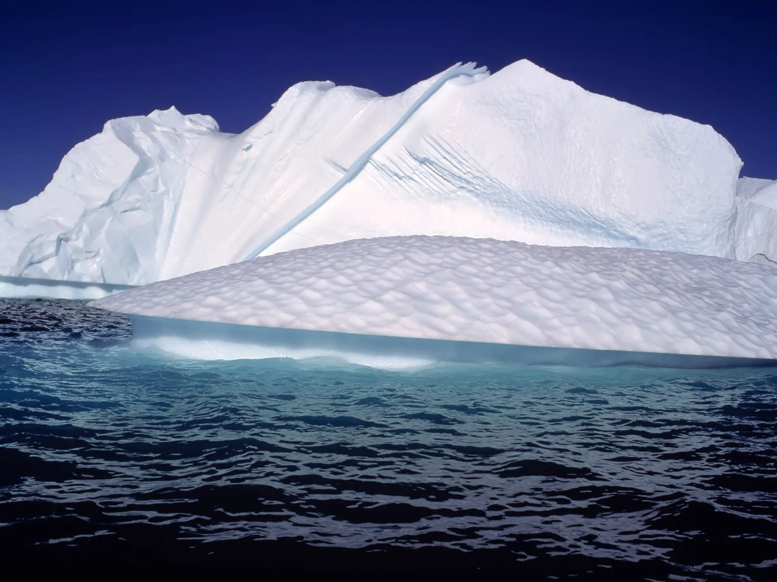 A large iceberg with a textured, layered shelf floats in the deep blue ocean under a clear sky.