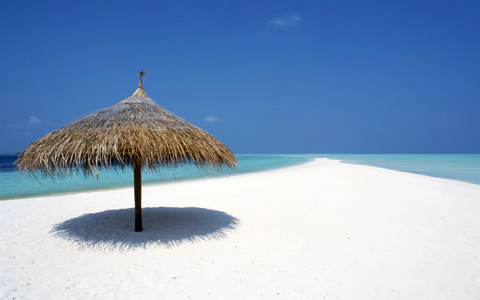 A solitary thatched umbrella casts a shadow on a pristine white sand beach.