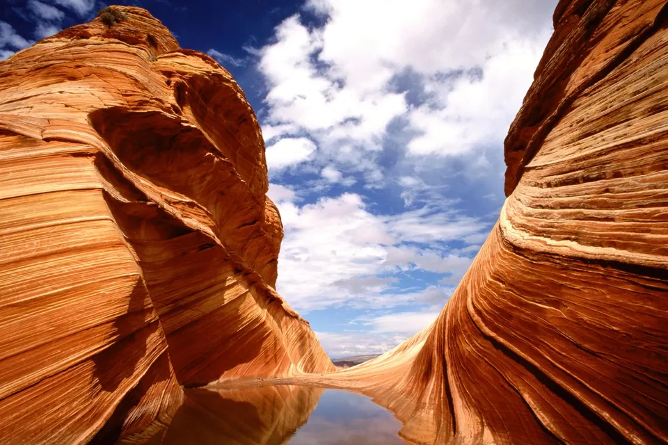 Swirling sandstone formations with a reflection pool under a blue sky with clouds.