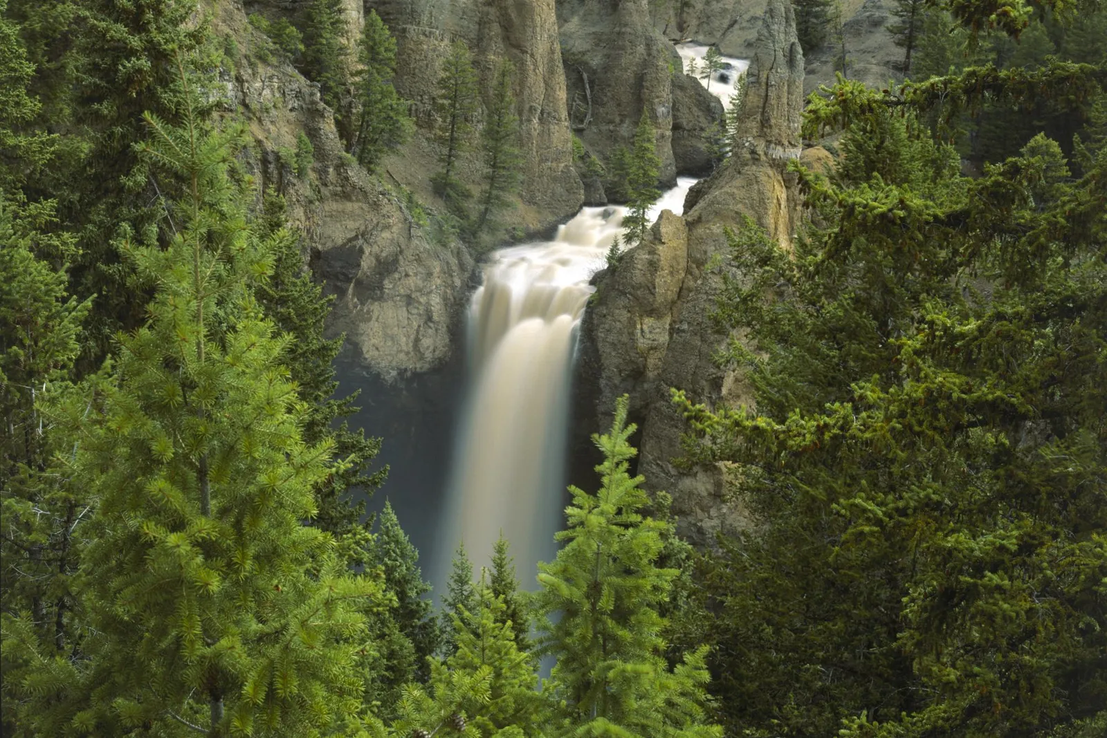 A tall waterfall plunges into a canyon, framed by evergreen trees and rocky cliffs.