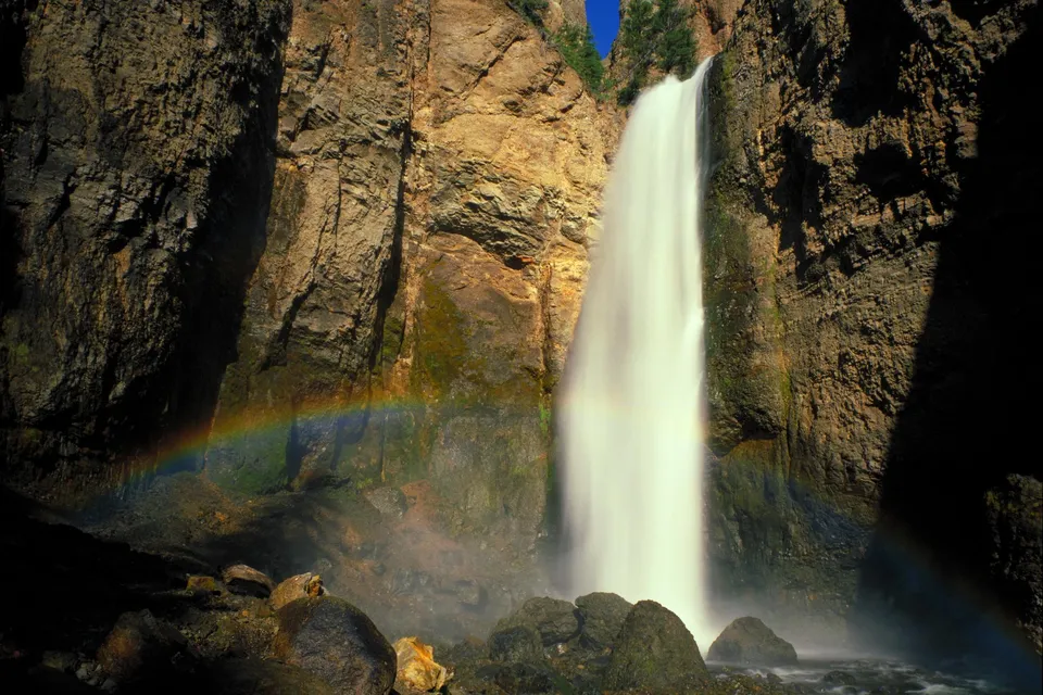 A vibrant rainbow arcs through the mist of a tall waterfall cascading down a rocky cliff face.