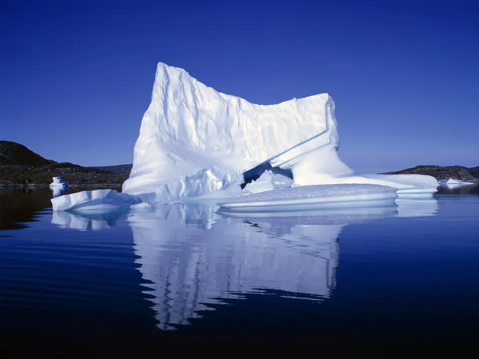 A large, textured iceberg stands tall and is reflected in the still, deep blue water under a clear blue sky.