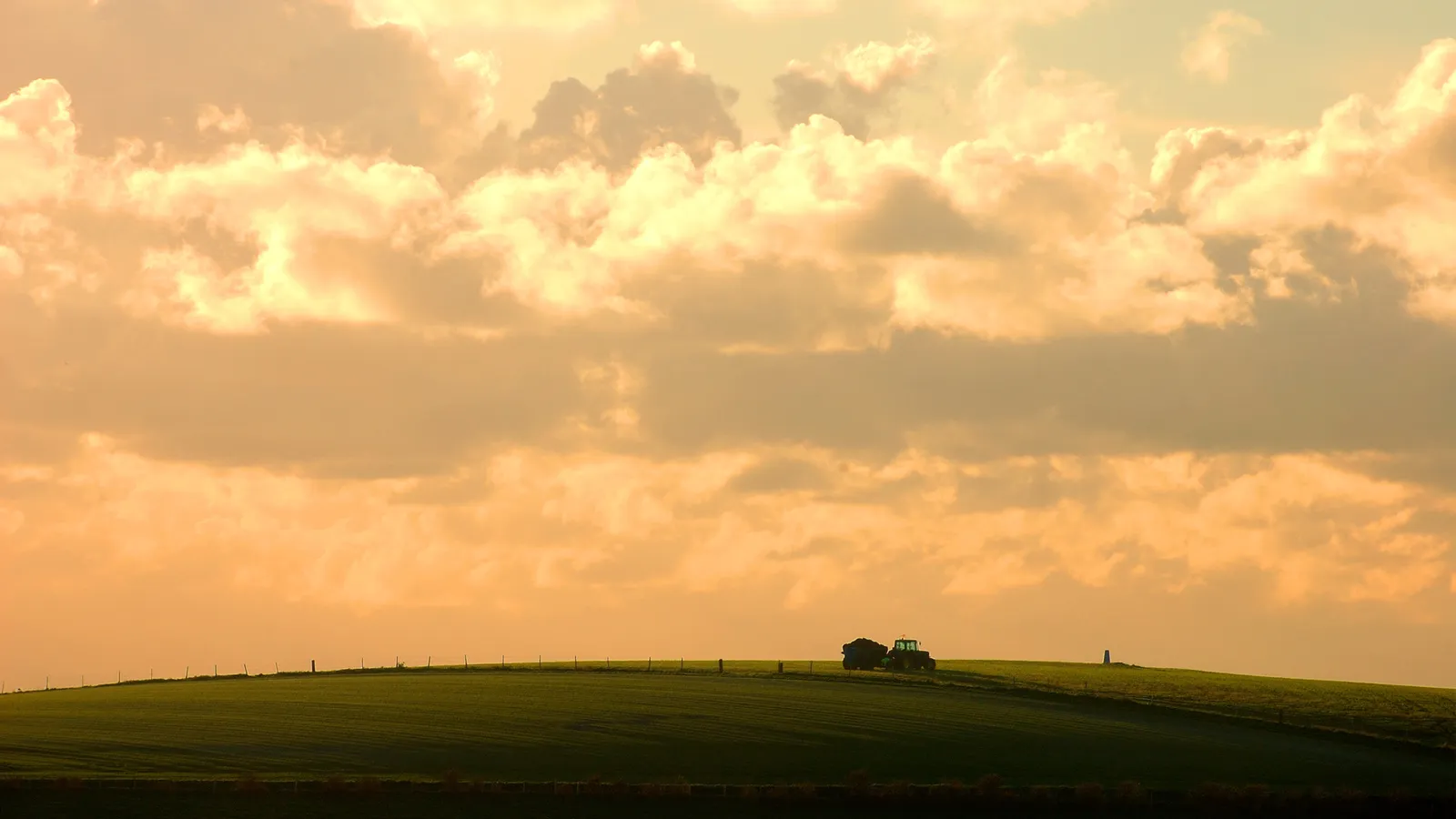 A silhouette of a tractor with a trailer on a green hill under a dramatic, cloudy sky.