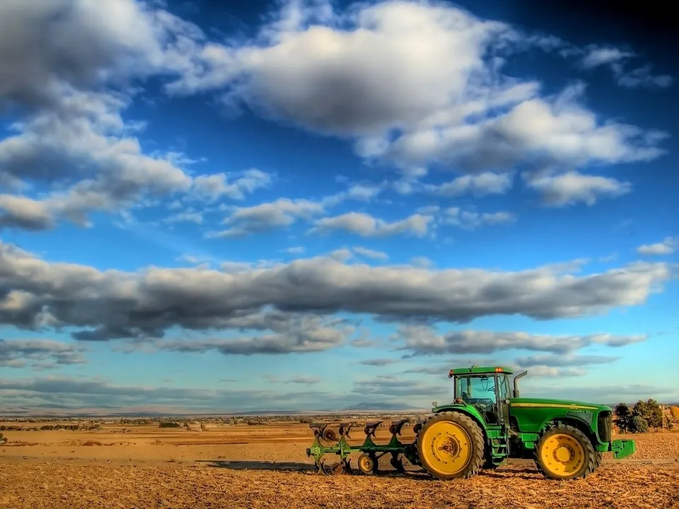 A green tractor with a plow attached sits in a dry, brown field under a blue sky with dramatic clouds.