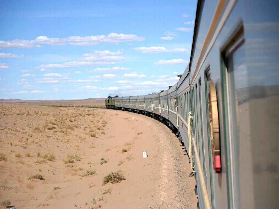 A long train travels through a vast, sandy desert under a bright blue sky with scattered clouds.
