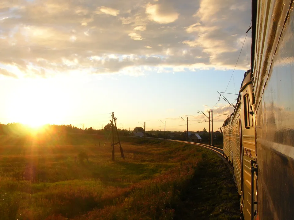A train travels along a track through a grassy field at sunset, with the sun low on the horizon.