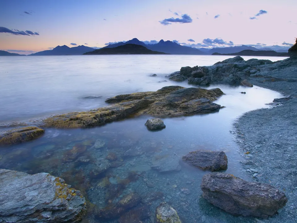 A serene lake shoreline with rocky outcrops and distant mountains under a twilight sky.