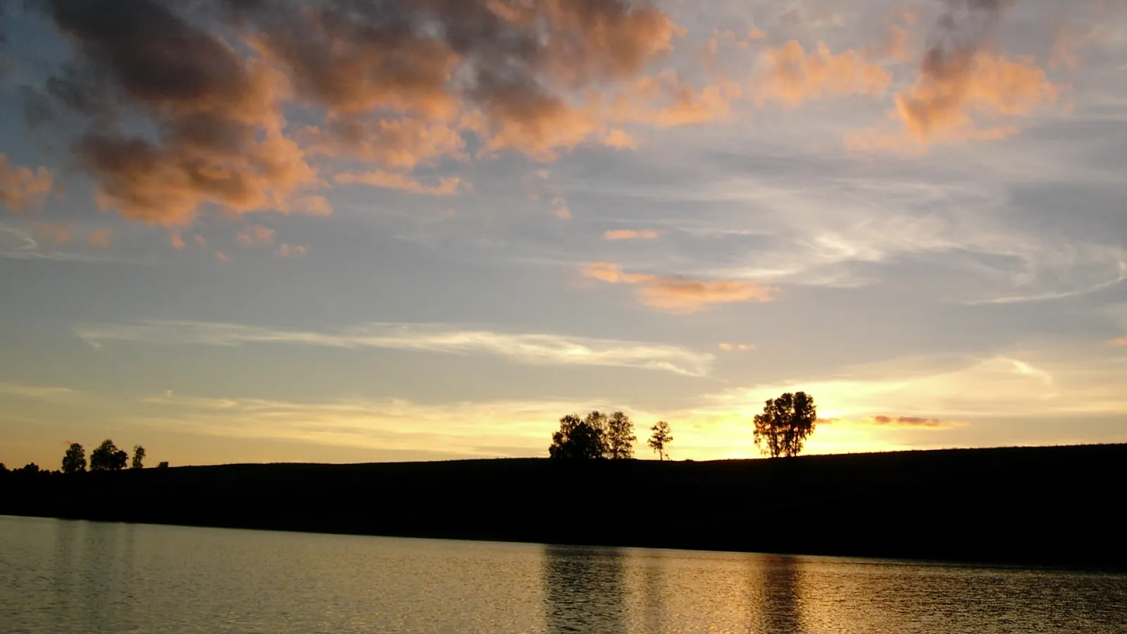 Silhouetted trees on the horizon reflect in a lake during a vibrant orange and pink sunset.