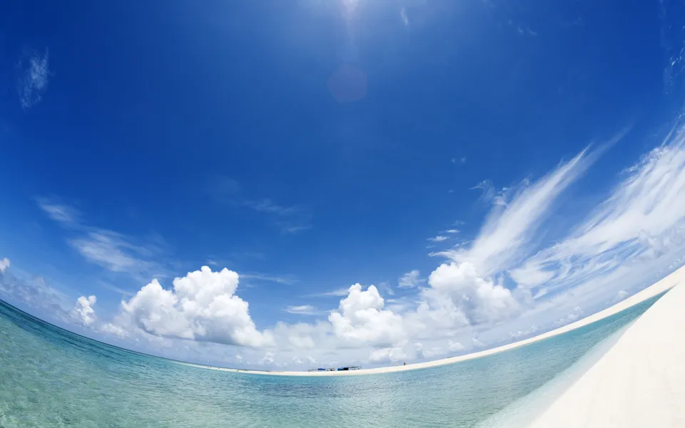 A wide-angle view of a tropical beach with clear turquoise water and a bright blue sky with scattered white clouds.
