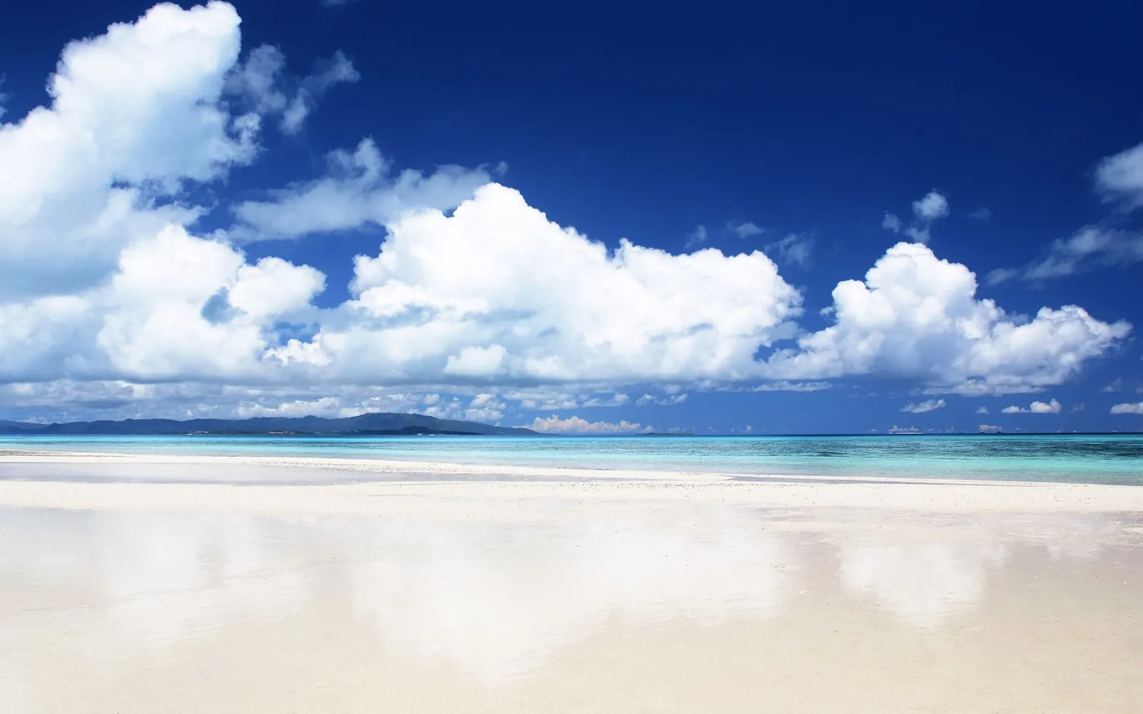 A wide, white sand beach meets turquoise water under a deep blue sky with fluffy white clouds.