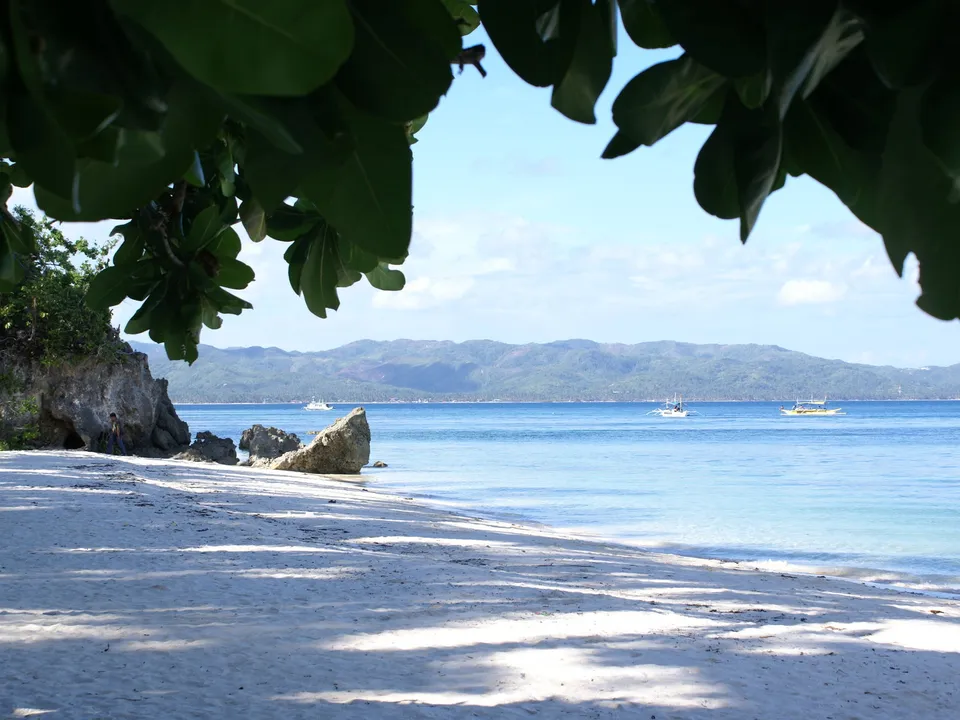 A white sand beach with clear blue water and distant boats, framed by lush green leaves.