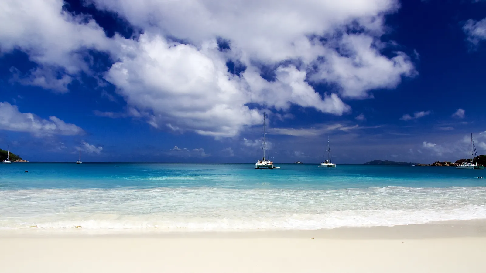 Several catamarans are anchored in a clear turquoise sea, with a white sand beach in the foreground and a blue sky with white clouds above.