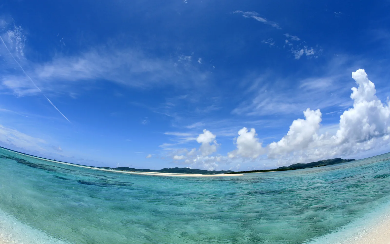 A wide-angle view of a tropical beach with clear turquoise water and a blue sky with scattered clouds.